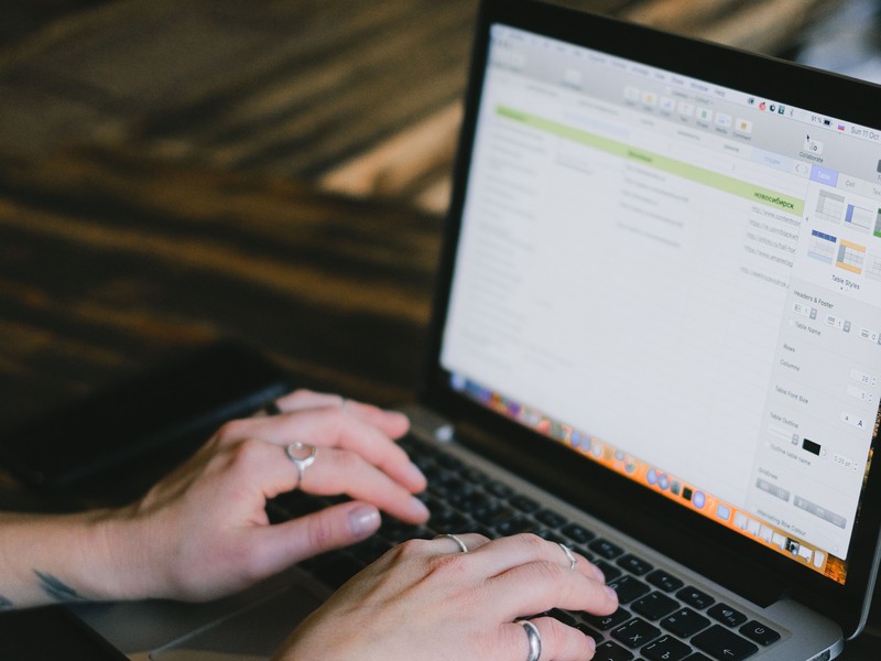Mãos com anéis digitando no teclado de um laptop aberto sobre uma mesa de madeira. A tela do laptop mostra um documento de texto ou planilha com informações e formatações.