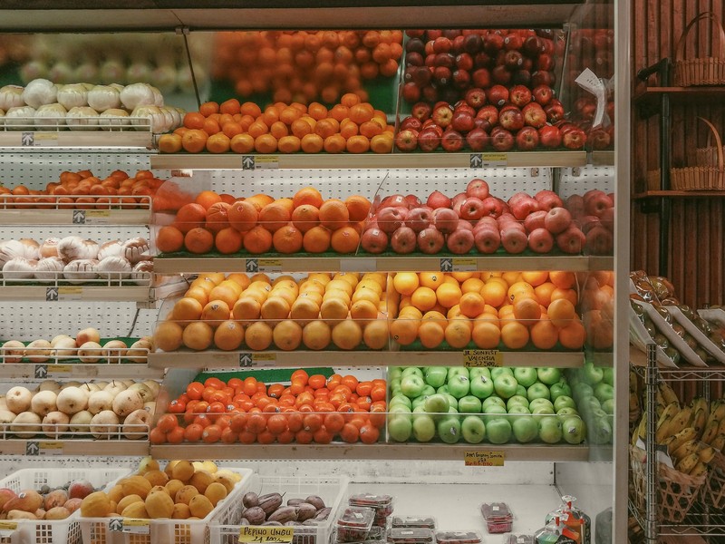Prateleiras refrigeradas de um mercado com frutas variadas organizadas por tipo, incluindo laranjas, maçãs vermelhas e verdes, tangerinas e cebolas, com etiquetas de preço visíveis.