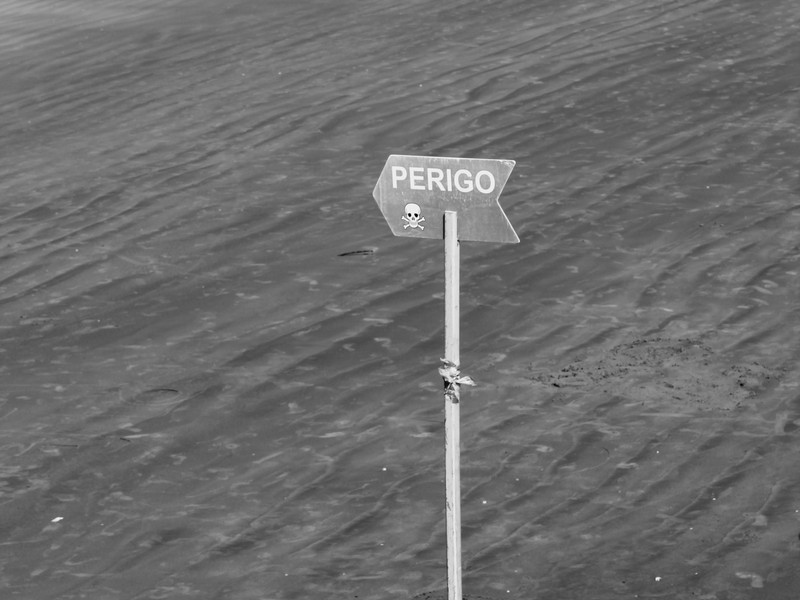 Fotografia em preto e branco de uma placa de madeira fincada na areia molhada com a palavra 'PERIGO' escrita em letras maiúsculas e o desenho de uma caveira com ossos cruzados logo abaixo.