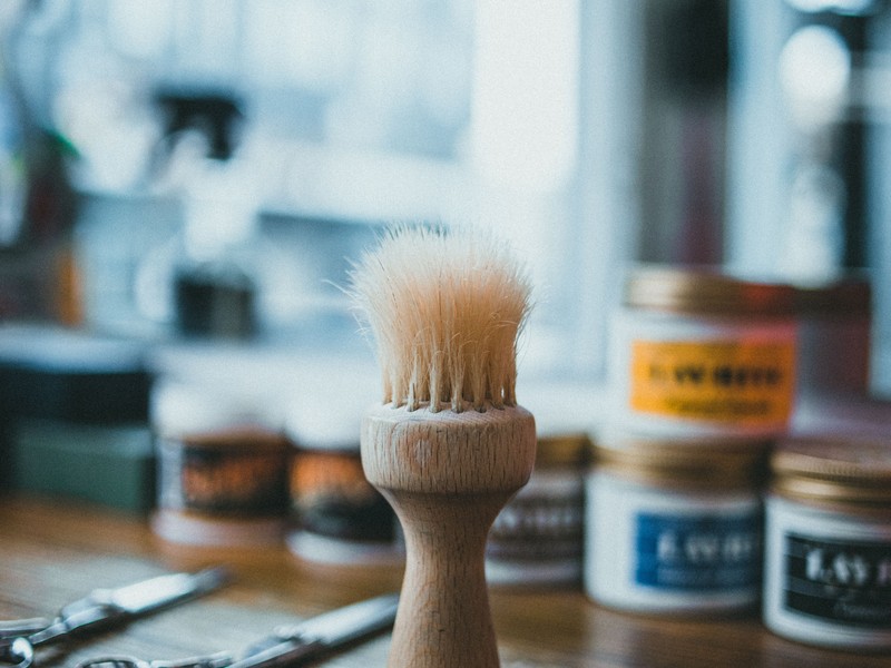 Pincel de barba de madeira com cerdas naturais sobre um balcão de barbearia, com tesouras e potes de produtos de barba desfocados ao fundo.